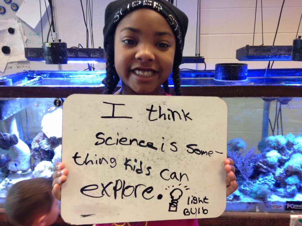 A young girl is asked to define science. She is standing in front of a fish tank holding a sign that says "science is something that kids can explore". Beneath the quote is a picture of a light bulb.