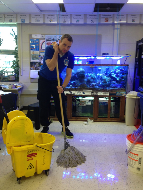 A male teacher is standing in front of an aquarium with a mop, looking tired. The teacher was working overtime to clean up spilled water caused by a student mistake. A float switch on the water reservoir solved this problem.